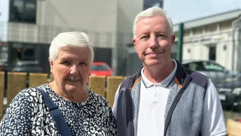 Jean has short grey hair and is wearing a navy and white printed top with a navy bag strap across her body. Next to her is Tom who has short grey hair and is wearing a white polo shirt, grey jumper and is smiling at the camera. Behind them is a brown wooden fence, a metal fence and a number of cars parked.
