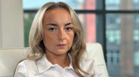 BBC Chloe Gibson is looking at the camera with a stern expression on her face. She is wearing a white shirt and has long blonde hair.