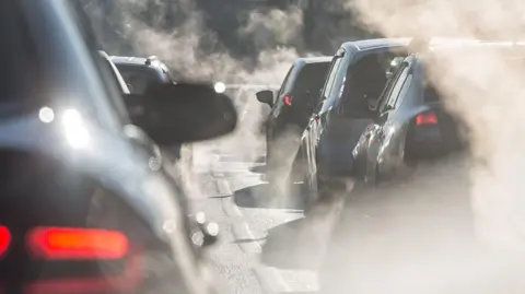 A number of cars waiting in traffic on a road. The cars are surrounded by exhaust emission. 