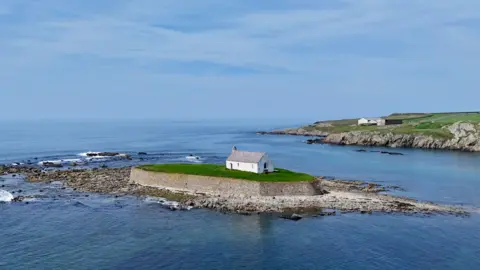 Visit Wales The image shows St Cwyfan's church. The church is small and white and on an elevated island surrounded by blue water, greenery and blue skies.
