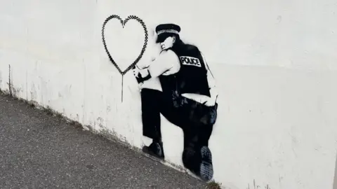 A spray painted stencil of a police officer kneeling down with a spray paint can which has drawn a love heart 