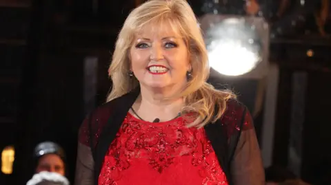 PA Linda Nolan is evicted from the Celebrity Big Brother House at Elstree Studios in Borehamwood in 2014. She has long blonde hair and is wearing a bright red dress with a black muslin cardigan
