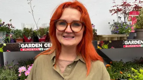 A woman with bright orange hair is smiling into the camera. She has a nose ring and is wearing large, brown glasses, a green short sleeved shirt and a green t-shirt. She is standing in front of a stall of flowers in plastic pots.