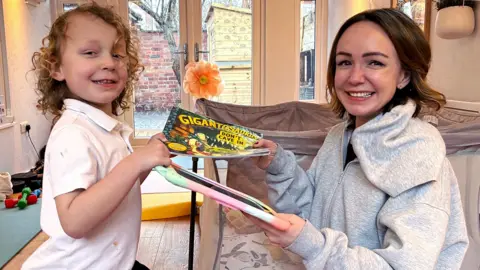 Romi, who has blond curly hair and is wearning a white t-shirt, is passing a book to his mum, Alexis who has short brown hair and is wearning a grey hoodie. She is passing a digital device with a colourful cover back to him and both are smiling. 