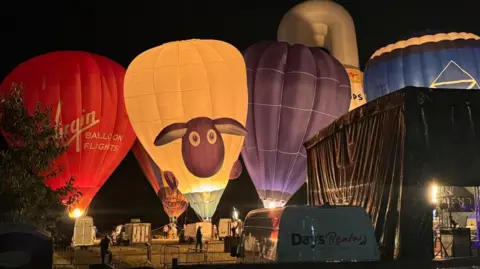 Four hot air balloons waiting to take off on a field at night time The balloon on the far right is red and has white writing that reads "Virgin Balloon Flights". Next to it is a white balloon with the face of a cartoon sheep on it, as well as a purple hot air balloon and a blue one. The sky is black in the background and the fire from the basket of the hot air balloons is illuminating the actual balloons.