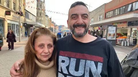 Henry Godfrey-Evans/BBC A man, Taskin Mehmet, and his mother, Layla Mehmet, stand in the high street. The man is wearing a black t-shirt and has a dark beard speckled with grey. The woman is in a brown rollneck jumper and has long, mid brown hair.
