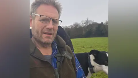 Steven Conisbee is wearing a brown gilet over a blue shirt with his black and white dog in the background in a selfie photograph. There are fields and woods behind him.