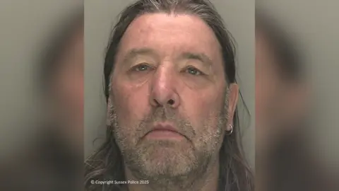 Sussex Police A mugshot of a man with an earring, grey stubble, and long hair.