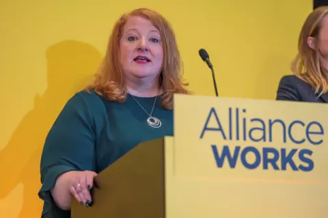 PA Media Naomi Long delivering a speech behind a yellow Alliance Works plinth