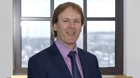 Close up of Mr Richard Hemans. He is standing in front of a building. Mr Hemans is wearing a navy suit with a purple shirt. 