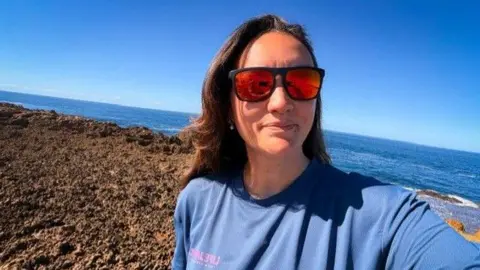 Lara Vafiadis Lara Vafiadis takes a selfie while wearing orange tinted sunglasses and a blue top. She has long brown hair and is standing in front of the ocean on a sunny and clear day.