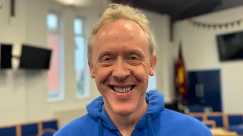 David Stanley is smiling inside a classroom. He is wearing a blue hoody.