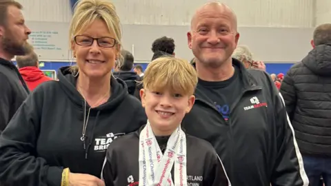Sharon Gill Theo with his mum (left) and dad (right) he is wearing medals around his neck. All three are smiling at the camera