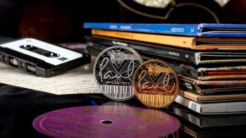 The Royal Mint A silver and gold coin upright, side by side, with Paul McCartney writing on, leaning next to a tape and books