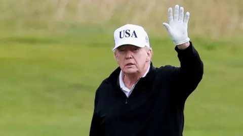 Reuters Donald Trump waves while playing on the Turnberry golf course. He is wearing black clothing and a white USA hat, with a white golf glove.