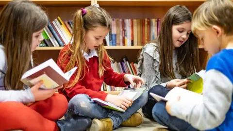 A group of children reading in a circle. Three girls and a boy are reading books with a book shelf behind them.