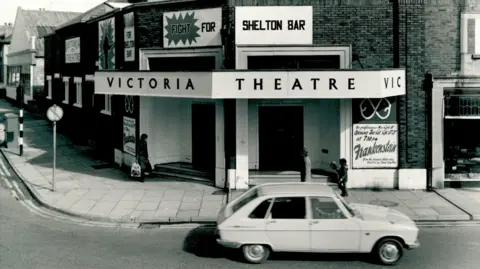 New Vic Theatre A black and white image of a brick built theatre with black lettering on a white canopy that read Victoria Theatre. There is a car parked at the front, and a pavement and road signs in front of the building.