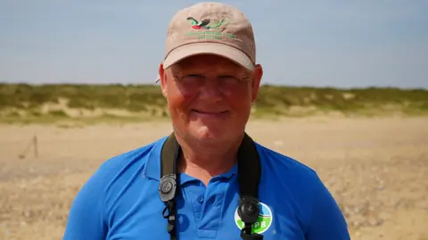 Shaun Whitmore/BBC A man smiling at the camera in a head and shoulders image. He is wearing a blue polo shirt and a beige baseball cap. 