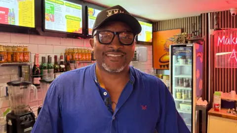 A man smiling and wearing a black hat with black glasses. He has a blue shirt on with a red logo. 