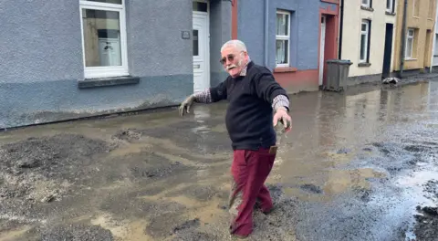 A man in burgundy trousers and a black jumper is wading through a muddy street with houses behind him 