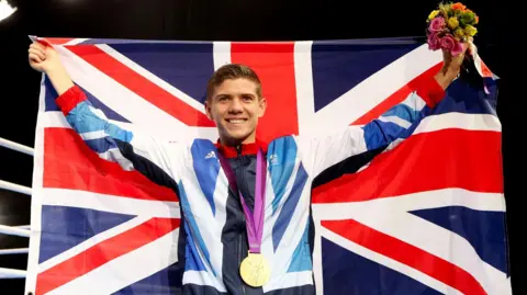 Getty Images Luke Campbell holds a Union Flag aloft as he smiles at the camera. He is wearing a Team GB tracksuit and has a gold medal around his neck.