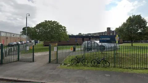 The outside of Lliswerry High School in Newport, south Wales. Green gates can be seen at the entrance alongside a navy school sign. A bike shed can be seen beyond the gates to the right of a path towards the school. Grass covered the ground around the path and two bikes lean against the gate. 