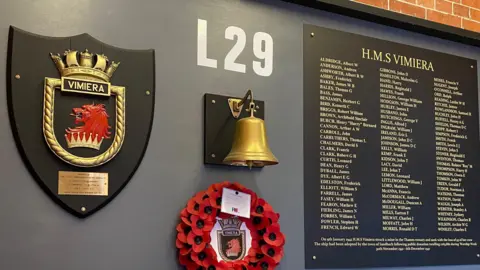 A large board mounted on a wall with a ceremonial shield on the left, a bell in the middle, a wreath underneath and a plaque on a right with HMS Vimiera at the top in gold letters and a list of 93 names.