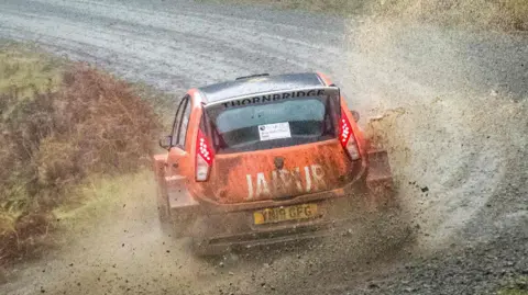 An orange rally race car travels at speed on a gravel track. The car has sponsorship stickers plastered across the side of the car, one reading 'JAIPUR'. The vehicle is travelling in a forest. 