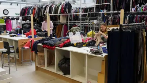 A man and a woman are in a large stockroom sorting through clothing with long tables and rails of clothes behind them.