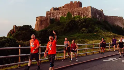 Two men in matching red T-shirts with logos on and a lanyard round their necks are taking part in the Around the Island Walk challenge. One is walking along a pavement and the other is next to him walking along the road. The man on the right has one arm in the air with his thumb pointing up. There are other race participants behind them.  