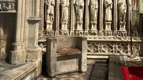 A squat stone stool, with a seat arms and low back, stands in a large minster church in front of a decorative screen featuring intricately carved stone statues.