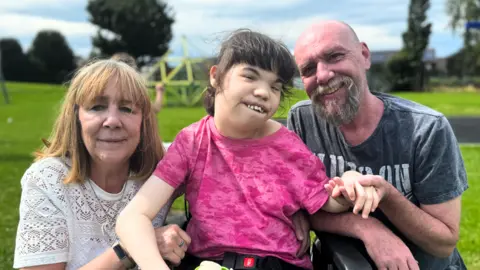 BBC Parents sit with their daughter who is in a wheelchair.