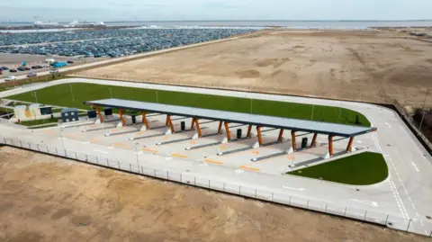 Milence An aerial adaptation of what the design of the HGV electric charging point would look like. A roof can be seen over eight charging ports with grass behind and a large car park next to it.