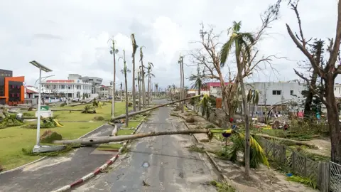AFP via Getty Images An aerial view of the city of Toamasina, on the east coast of Madagascar. A road lined with palm trees heavily damaged by Cyclone Gezani, with several trees snapped or uprooted and lying across the street. Debris and fallen branches cover the area, surrounded by buildings, under a cloudy sky.