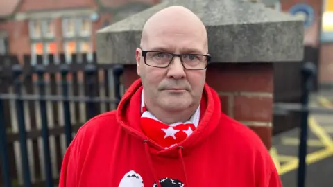 A man standing in front of a black fence. He is wearing a red hoodie with a red and white knitted scarf tucked into it.