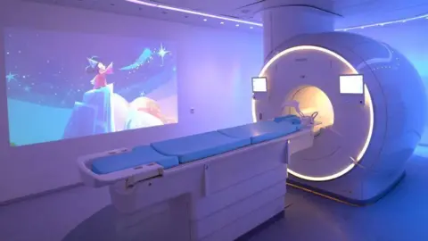 Raoul Hollants An MRI scanning suite bathed in a lilac-coloured light with a cartoon image of Mickey Mouse projected on to the wall next to the scanner.