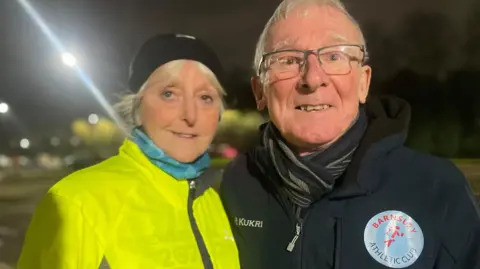 Simon Thake An elderly couple in bright running jackets and hats huddle together, smiling.