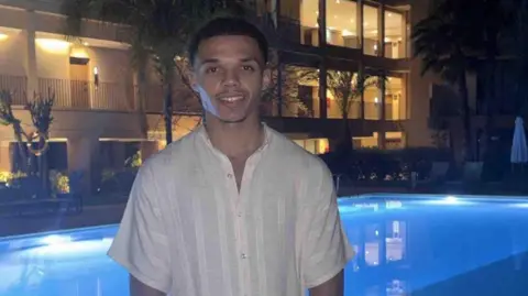 Sam Rushton Young man with short hair wears white shirt and stands posing for a photo in the evening with a pool and hotel rooms in the background