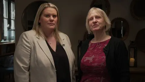 BBC Micala and Sue stand side by side looking defiant. Micala has long blond hair and is wearing a black shirt and light brown jacket. Sue has short blond hair and is wearing a red top with brown horses on it and a black jacket. Behind them is a wall with several mirrors on it 