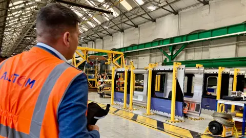 A man in a blue jacket and orange hi vis vest is standing in a factory beside a train.