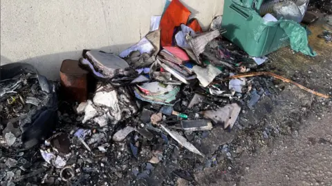 A pile of burned objects on the street outside the fire damaged home. A melted green recycling box is visible to the right and only a few books can be made out amongst the pile of black and melted waste