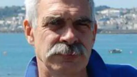 BBC A picture of a man smiling at a camera. He has grey hair and grey moustache. He is wearing a blue shirt. 