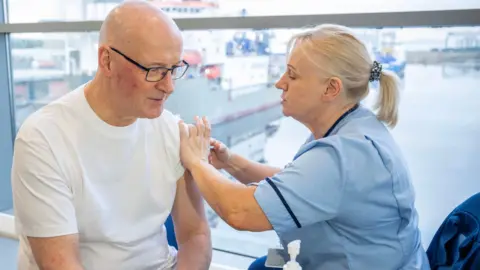 PA Media John Swinney, a bald man with glasses, gets his flu jab from a blond-haired nurse wearing a light blue uniform. He is wearing a white t-shirt.