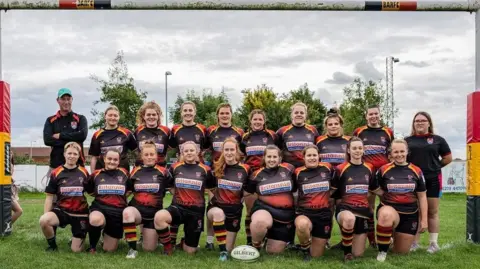 Bridgwater and Albion Rugby Club Two rows of rugby players stood under the posts with coaches either end of the rows. Their kit is black, red and yellow. A white rugby ball is on the grass in front of them