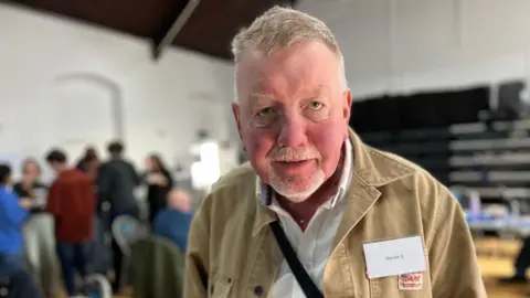 BBC/Richard Moss Steven Shepherd at Seaton Delaval arts centre. He has short white hair and a goatee and blue eyes. He is wearing a white shirt and beige denim jacket. He has a name tag pinned to it. He is looking at the camera with a serious expression and his mouth slightly open.