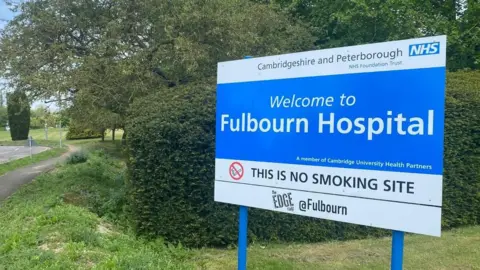 A blue and white sign saying Cambridgeshire and Peterborough NHS Foundation Trust, and Welcome to Fulbourn Hospital, standing on blue legs, against a dark green hedge, on a lighter green verge beside a road