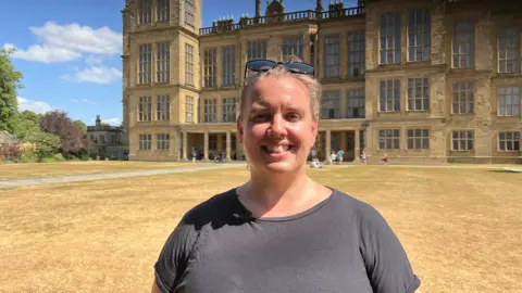 Emily Eastwood smiling and wearing a dark grey top, stood in front of Hardwick Hall, a grand three story building.