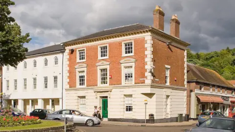 The outside of the headquarters of Hampshire Community Bank - a three-storey brick building in a residential area. It has two chimneys and a green door.