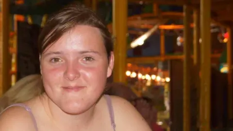 Supplied A woman is looking into the camera and smiling. She has light brown hair in a ponytail and is wearing a light purple top with straps around her shoulders.