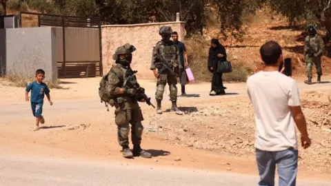 Una toma panorámica muestra a tres soldados de las FDI con uniforme militar, cubrebocas y fusiles, caminando por un camino polvoriento. Cuatro residentes de Cisjordania (dos hombres, una mujer y un niño) se ven caminando alrededor de los soldados. Uno de ellos sostiene un teléfono inteligente frente a su rostro para grabar o tomar una foto.